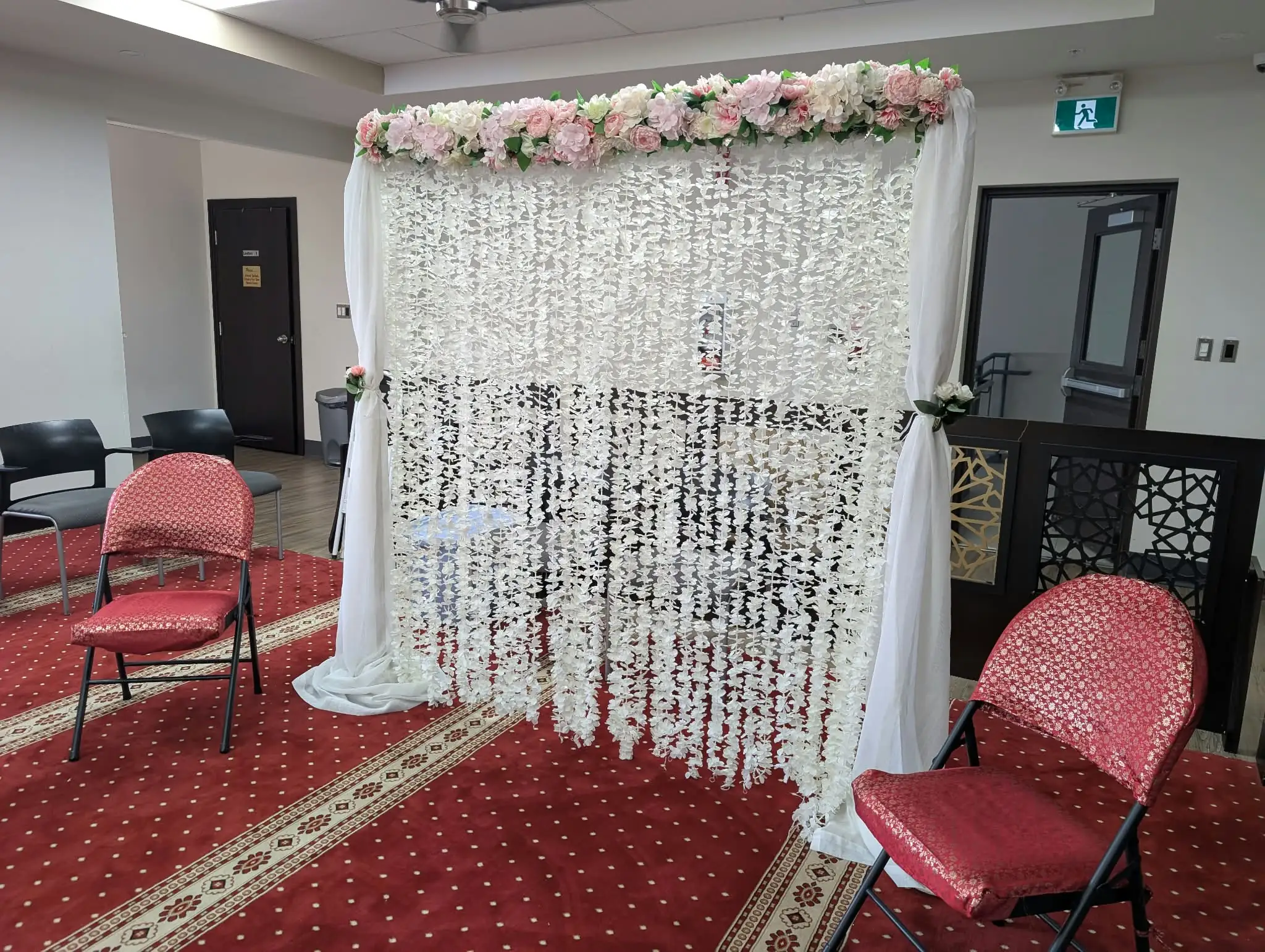 The see-through flower wall with a pink and ivory floral header and chiffon side curtains, set up at Jame Masjid in Mississauga between two chairs